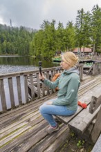 Woman sitting on a wooden footbridge and holding a camera over a lake, Mummelsee, Seebach, Ortenau