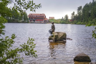 A statue sits on a rock in the lake, with a building in the background, Mummelsee, Seebach, Ortenau