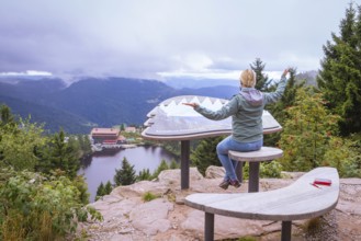 Woman sitting on a wooden bench at a viewpoint, overlooking a lake and mountains, Mummelsee,