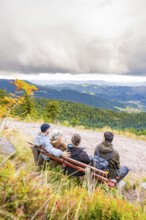 Three people on a bench enjoy the wide view over wooded mountains and valleys, Mummelsee, Seebach,