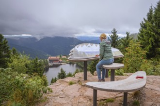 Woman at the panoramic viewpoint with information board, view of lake and forest under clouds,
