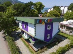 An upside down house with green and purple colours in a park surrounded by trees, aerial view, Ters
