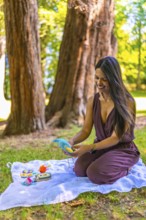 Happy young latin mother kneeling on a blanket in a park, preparing vibrant baby toys while