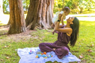 Young latin mother playing with her baby girl in a park, lifting her in the air and smiling,