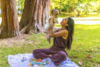 Latin mother joyfully lifting her baby daughter while sitting on a blanket in a park, enjoying