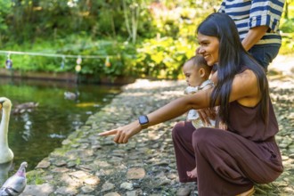 Mother and baby daughter pointing at birds by the pond in a botanical garden, enjoying a sunny day