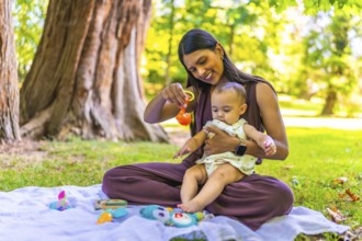 Latin mother holding her baby daughter and playing with colorful toys while sitting on a blanket in