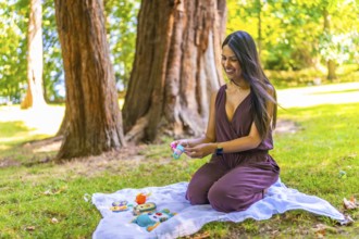 Smiling latin mother arranging baby toys on a blanket in a park, enjoying a sunny day outdoors