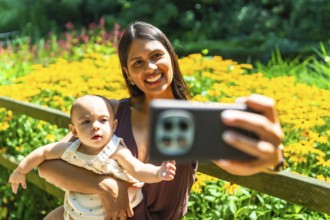 Happy latin mother holding her baby daughter, taking a selfie with a smartphone in a garden full of