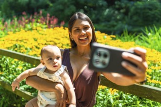 Happy latin mother holding her baby daughter and taking a selfie with smartphone in a beautiful