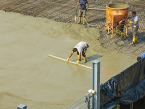 Workers smoothing concrete on a construction site while a concrete mixer stands in the background,