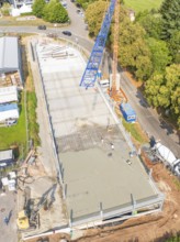 Top view of a construction site with crane and workers, surrounded by trees and roads, car park