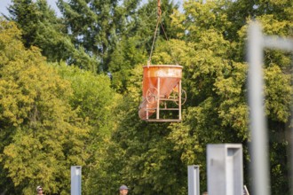 An orange-coloured concrete bucket hovers over a building site. Trees and a clear sky in the