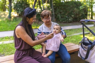 Mother feeding her baby girl solid food, with grandmother holding her, sitting on a park bench