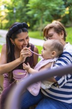 Mother feeding her baby a meal in the park, with grandmother holding the child, enjoying family