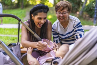 Latin mother feeding her baby girl solid food from a pink bowl, with grandmother assisting, in