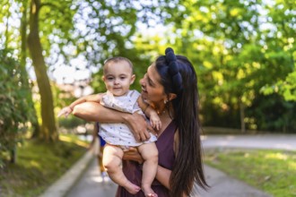 Happy latin mother smiling while holding her baby girl in a park, enjoying a sunny summer day