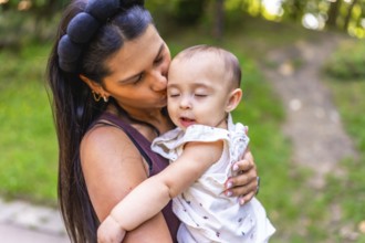 Young latin mother kissing her baby daughter while holding her lovingly in her arms, surrounded by
