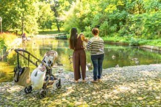 Mother, grandmother and baby girl enjoying a sunny day by the pond, watching ducks swim in a park