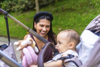 Happy latin mother entertaining her baby sitting in stroller at a park, enjoying family time