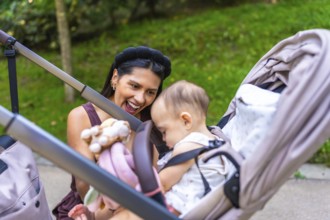 Happy latin mother showing toys to her baby daughter sitting in a stroller, enjoying a sunny day
