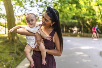 Happy latin mother holding and playing with her baby daughter in a park, enjoying a sunny day