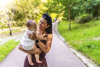 Latin mother holding her baby daughter, smiling and enjoying a walk in a park during a sunny day