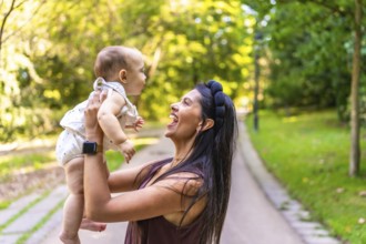 Smiling latin mother lifting her baby daughter while playing together in a sunny park, enjoying a