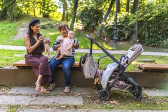 Grandmother holding baby girl while mother is eating and stroller is parked next to them on bench