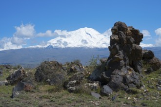 Snow-covered mountain under blue sky with clouds, surrounded by rocks and green landscape, aerial