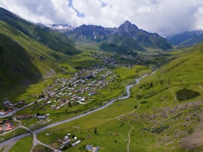 Wide valley with river and village, surrounded by mountains under a partly cloudy sky, aerial view,