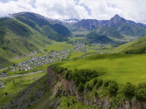 Lush valley with small village and rock face, framed by forested mountains under clouds, aerial