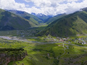 Green valley with river and village, surrounded by high mountains and cloudy sky, aerial view, view