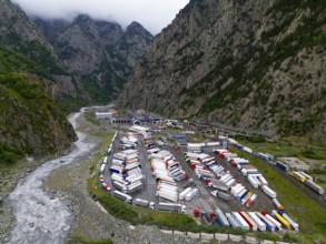 Car park full of lorries in a narrow mountain valley next to a river under a cloudy sky, aerial