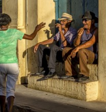Two old Cubans smoke cigars and sit on a staircase in the warm light of the sunset, they greet a