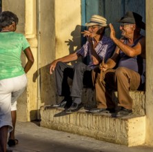Two old Cubans smoke cigars and sit on a staircase in the warm light of the sunset, they greet a