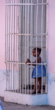 Little coloured girl behind window bars in a pink house, street of Trinidad, Cuba, Sancti Spíritus