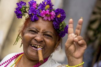 Peace sign, old Cuban woman with flowers and cigar posing for tourists for a tip, La Habana,