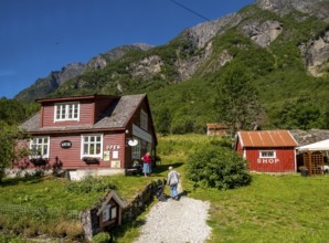 Tourists at mountain hut station, red wooden houses, gravel path, mountains, meadow, blue sky,