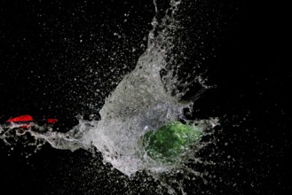A green water balloon bursts after arrow penetration, causing water to splash, in front of a dark