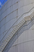 A long metal ladder stretches along a large cylinder building with shadows in the background,