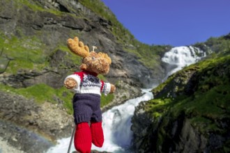 Kjosfossen waterfall, mascot moose in front of waterfall, mountain, rock faces, meadow, sky,