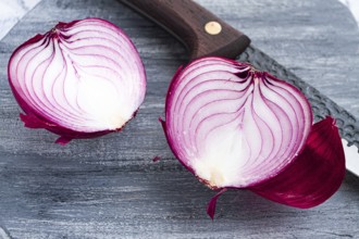 A vibrant red onion is cut in half, on a rustic wooden chopping board beside a sharp knife, ready