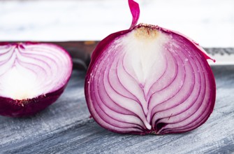A vibrant red onion is cut in half, on a rustic wooden chopping board beside a sharp knife, ready