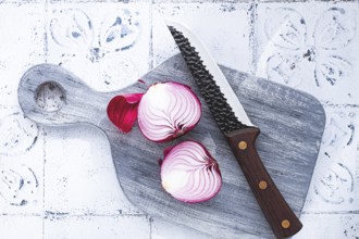 A vibrant red onion is cut in half, on a rustic wooden chopping board beside a sharp knife, ready