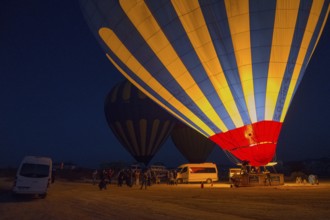 Hot air balloons at night with people and vehicles, bright colours in contrast to the dark sky,