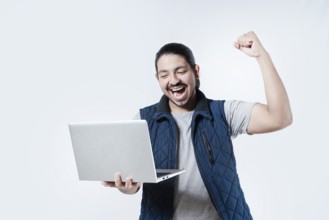 Young man celebrating holding a computer on isolated background. Male celebrating with fist in the