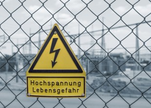 Yellow german high voltage warning sign on a mesh wire fence in front of a blue toned electrical