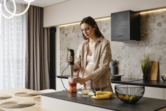 In a stylish kitchen, a pregnant woman blends fresh fruits like bananas and strawberries. Sunlight