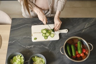 A pregnant woman with long hair skillfully slices cucumbers on a cutting board in a stylish kitchen